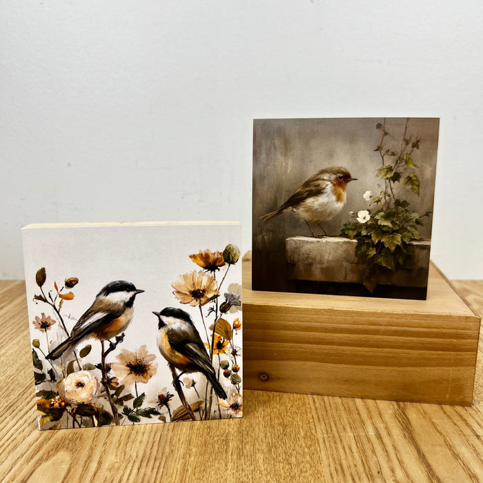 Two square art prints displayed on a wooden block and tabletop. One print shows two birds perched among flowers, and the other shows a small bird beside a stone planter with flowers.