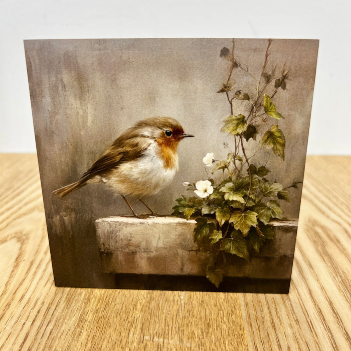A square art print or card featuring a small robin bird perched beside a stone ledge with white flowers and green leaves, displayed on a light wood surface against a plain light background.