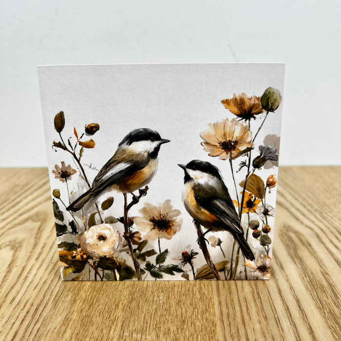 A square art print or card featuring two birds perched among delicate flowers, displayed upright on a light wood surface against a plain light background.