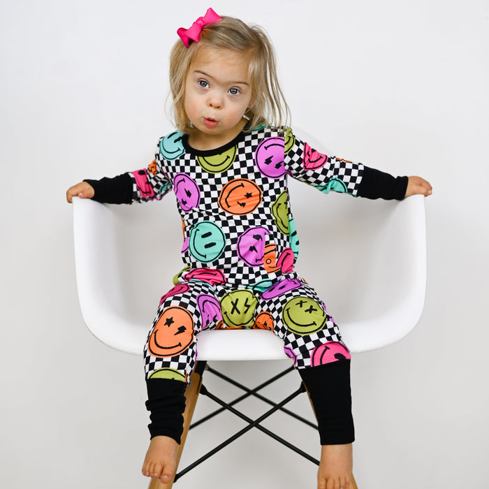 Child wearing a colorful checkered outfit with smiley face patterns on a white chair.