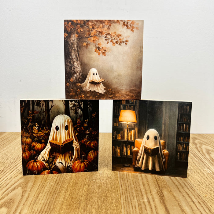 Three vertical art prints or cards displayed on a light wood surface against a white wall. Each print shows a ghost-like figure reading a book in different settings: one under an autumn tree, one among pumpkins in a dark forest, and one in a cozy book-filled room with a lamp.