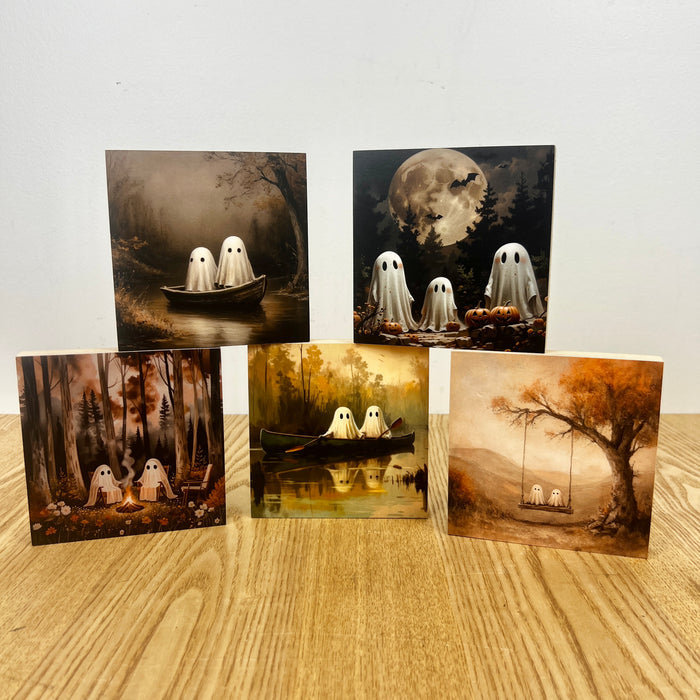 Five square Halloween-themed art prints or cards arranged on a light wood surface against a plain white wall. The designs feature ghost figures in a boat, ghosts in a forest, ghosts near pumpkins under a full moon, ghosts by a lake, and ghosts on a swing in an autumn tree.