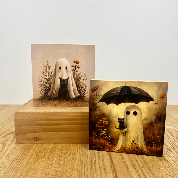 Two square art prints displayed on a wooden surface against a white wall. One print shows a white ghost with a black cat in a sepia-toned field.