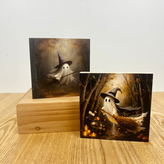 Two square art prints featuring a whimsical ghost in a witch hat riding a broomstick in a dark forest, displayed on a light wood surface with one print standing on a wooden block.