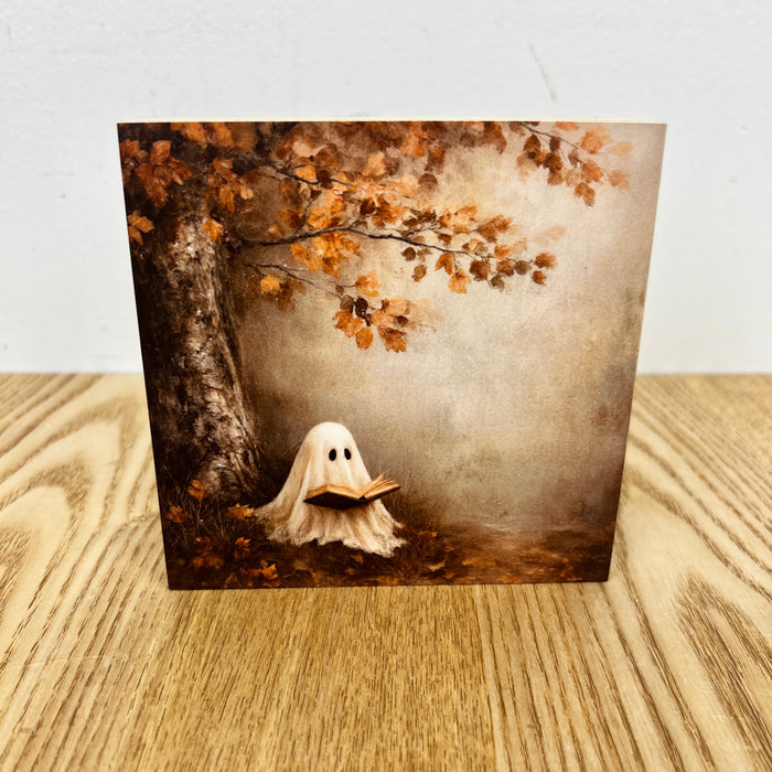 A square art print or card featuring a whimsical scene of a small white ghost reading under an autumn tree with brown leaves, displayed on a light wood surface against a white wall.