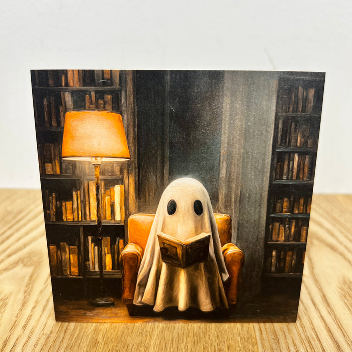 A square art print or card featuring a whimsical illustration of a ghost-like figure reading a book in a cozy armchair, surrounded by bookshelves and a lit lamp, displayed on a light wood surface against a white background.