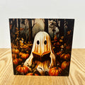 A square art print or card featuring a whimsical illustration of a ghost-like figure in a white sheet reading a book, surrounded by pumpkins in a dark forest, displayed on a light wood surface against a white wall.