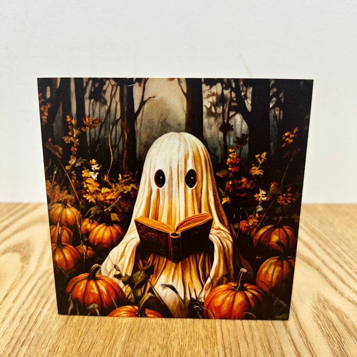 A square art print or card featuring a whimsical illustration of a ghost-like figure in a white sheet reading a book, surrounded by pumpkins in a dark forest, displayed on a light wood surface against a white wall.