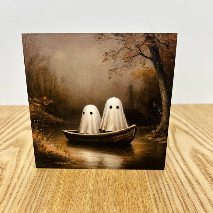 A square art print or card showing two white ghost figures with black eyes sitting in a small boat on a dark river, photographed standing on a light wood surface against a white wall.