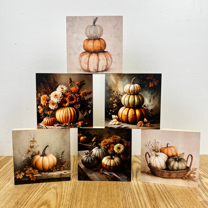 Five square art prints featuring autumn pumpkins and floral still life scenes, arranged on a light wood surface against a plain off-white wall.