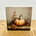 A square art print featuring a rustic still life of a light orange pumpkin surrounded by autumn leaves, displayed on a light wood surface against a white wall.